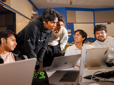 A group of five students lean over laptops in a classroom